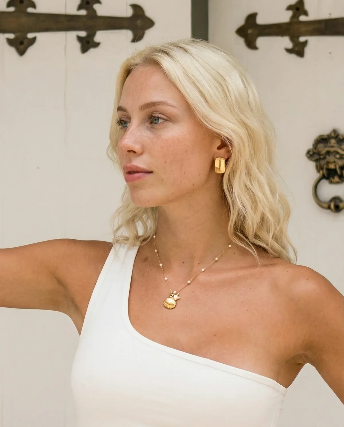 Blonde model in a one-shoulder top wearing a gold shell pendant and chunky gold earrings