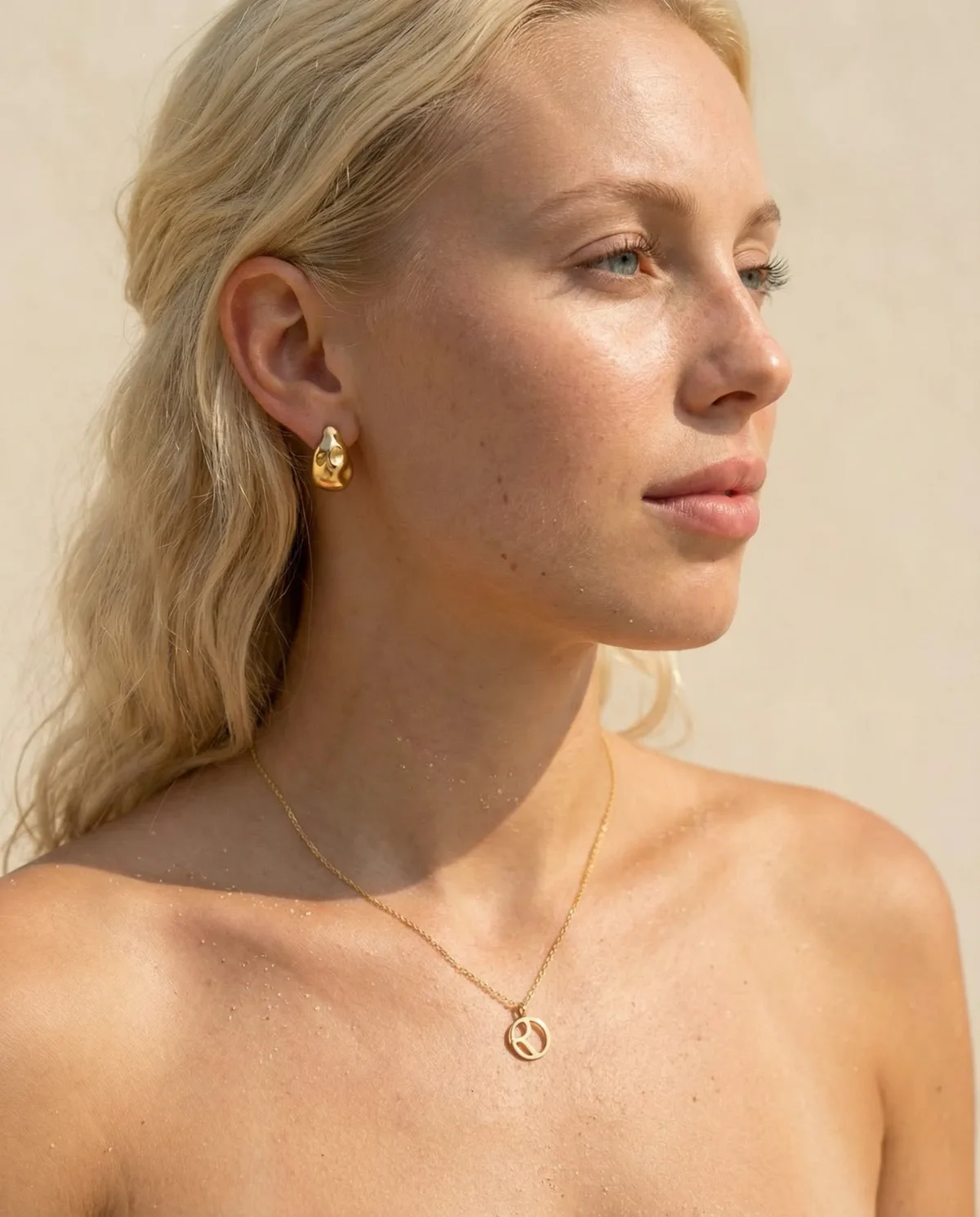 Blonde model in warm sunlight wearing a sculpted gold earring and initial pendant
